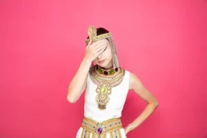 Beautiful woman like cleopatra in ancient egyptian costume looking tired and bored covering eyes with palm on pink