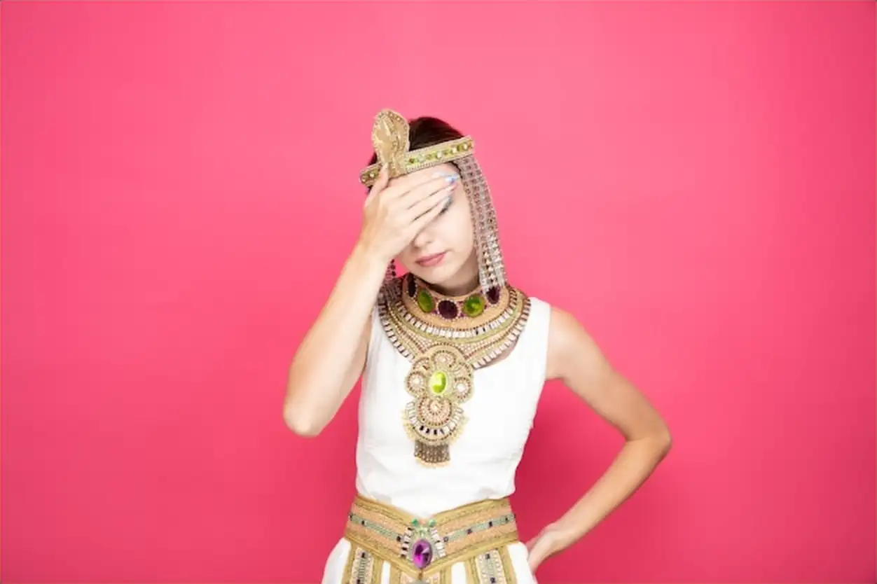 Beautiful woman like cleopatra in ancient egyptian costume looking tired and bored covering eyes with palm on pink